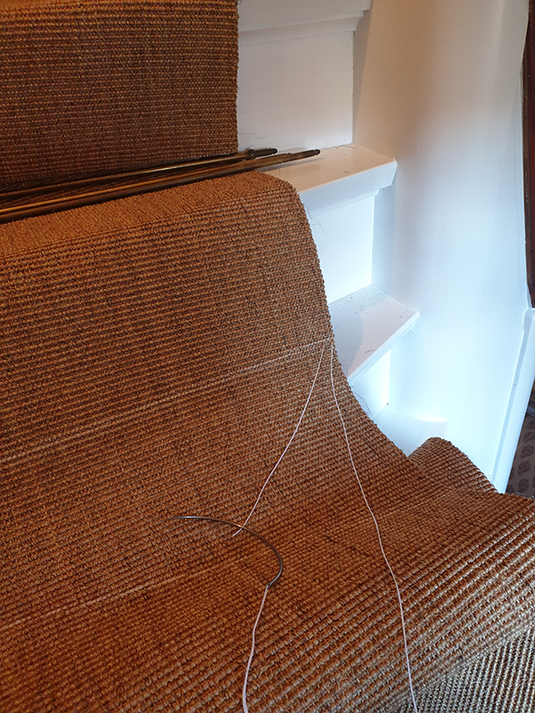 Brown textured fabric stretched over furniture with a sewing needle and white thread lying on it, near white stairs in the background.