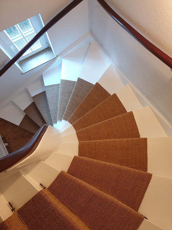 Spiral staircase with white steps and brown carpet runners viewed from the top near a window.