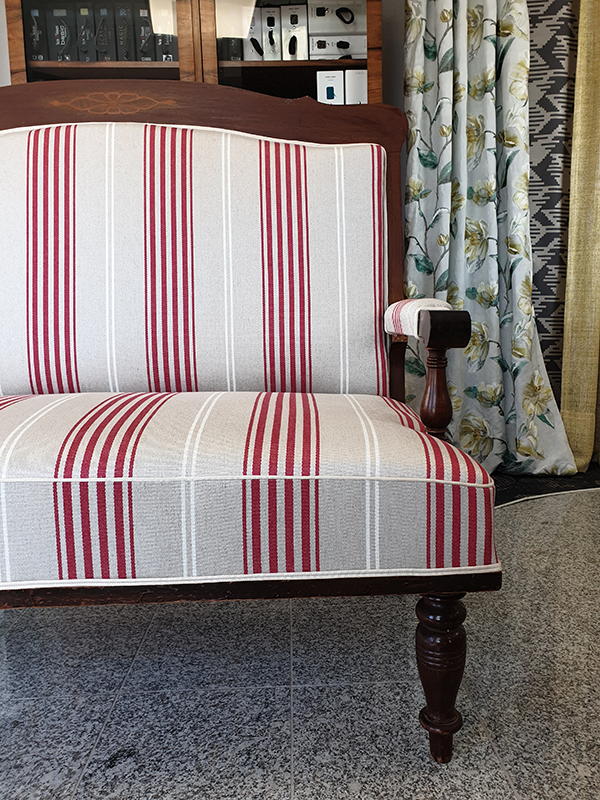 Wooden framed armchair with beige upholstery featuring vertical red and white stripes, placed on a tiled floor with patterned curtains in the background.