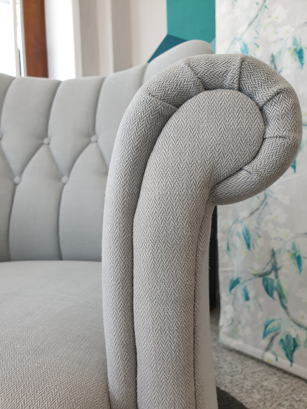 Close-up of a light gray upholstered sofa armrest with a rolled design and button-tufted backrest.