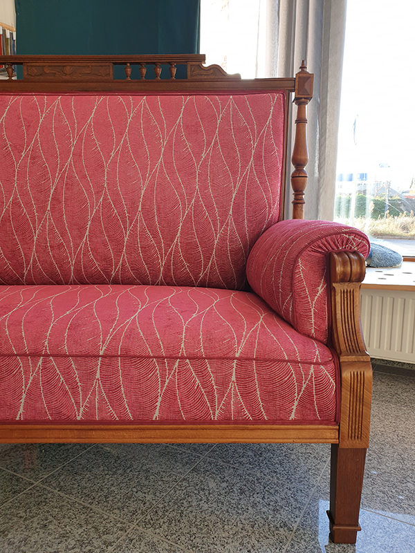 Vintage wooden armchair with red patterned upholstery and curved armrest, placed on tiled floor near a window.