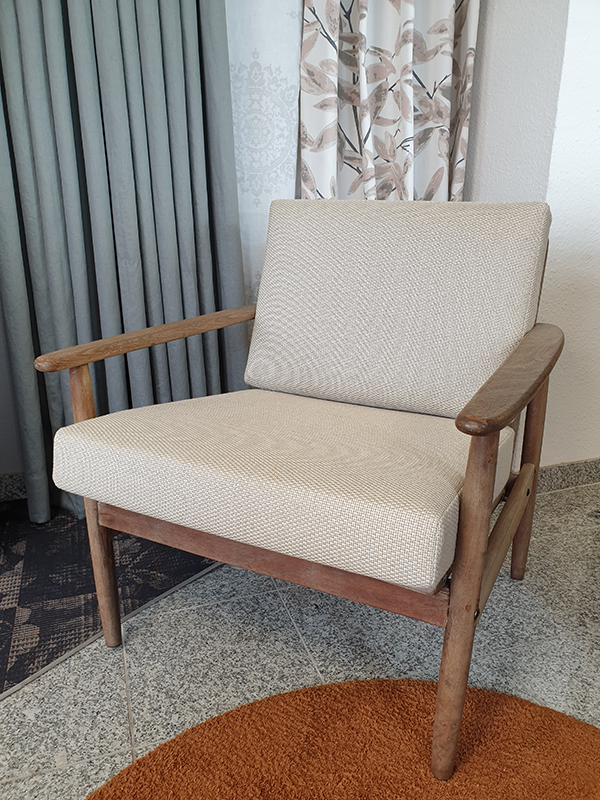 Beige cushioned wooden armchair placed on a tiled floor with a round orange rug in front and curtains in the background.