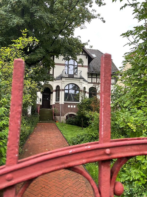 Two-story house with brick and white exterior, seen through a red gate, surrounded by greenery.