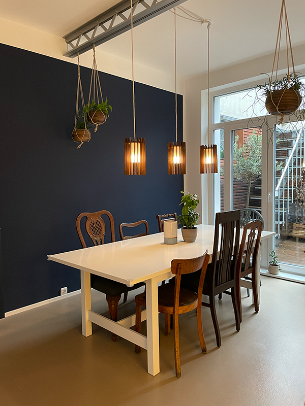 Dining area with white table surrounded by six wooden chairs, three pendant lights hanging above, and hanging potted plants near a large window.
