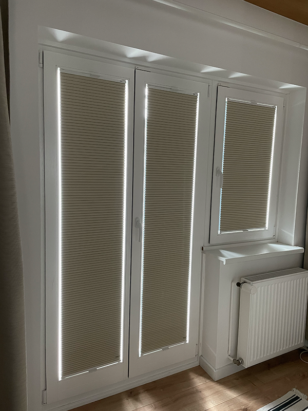 Three white-framed windows with beige pleated blinds partially closed, next to a white radiator on a wooden floor.