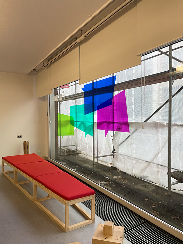 Indoor space with a long wooden bench with red cushions next to a large window adorned with colorful sticky notes in blue, green, and purple.