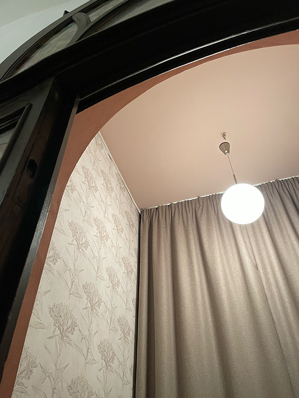 View of a ceiling with a round white pendant light, beige floral wallpaper on the left wall, and closed gray curtains on the right.