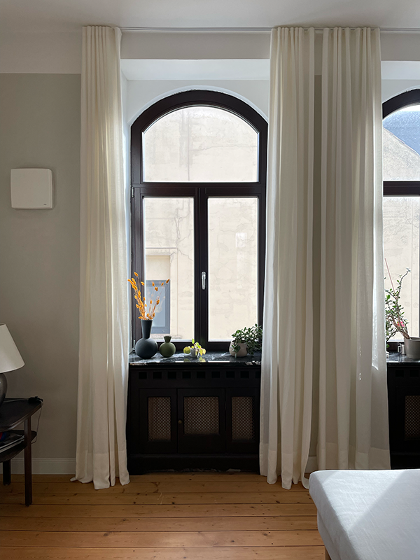 Arched window with black frame and white curtains flanked by a wooden floor and a white bed corner, featuring vases and plants on the window sill.