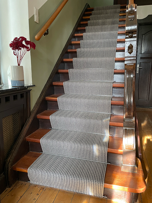 Top view of wooden staircase with patterned black and white runners on each step, a wooden handrail, and surrounding wooden floor flooring.