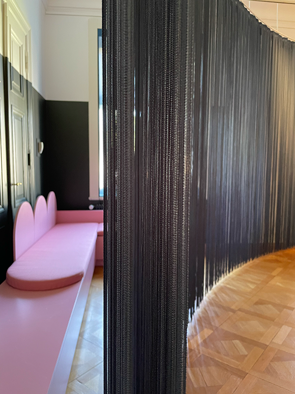 Curved room divider made of thin black hanging strings next to a pink bench with rounded backrests in a room with wooden parquet flooring.