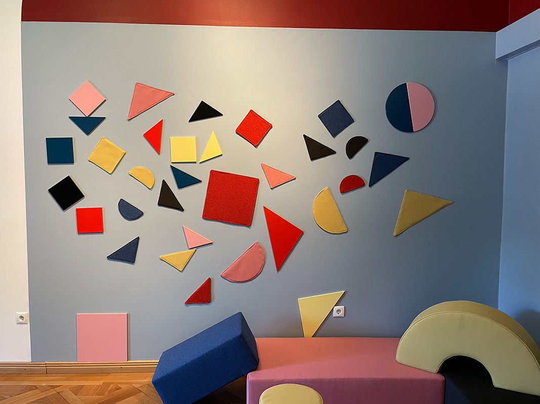 Wall decorated with various colorful geometric shapes including squares, triangles, and semicircles, with cushioned seating in blue, pink, and yellow underneath.