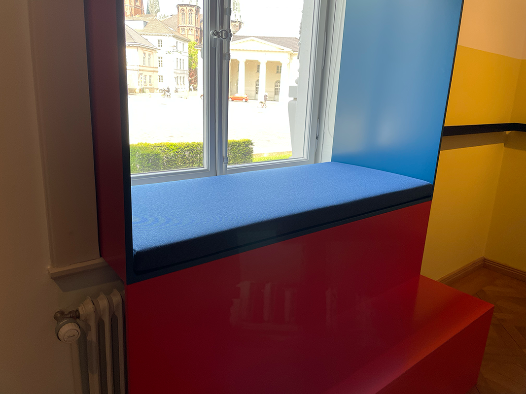 Blue cushioned window seat framed by red and blue panels inside a room with a radiator and wooden parquet floor.