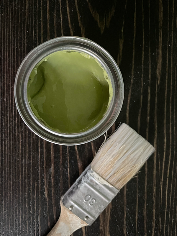 Open can of green paint next to a worn paintbrush with white bristles on a dark wooden surface.