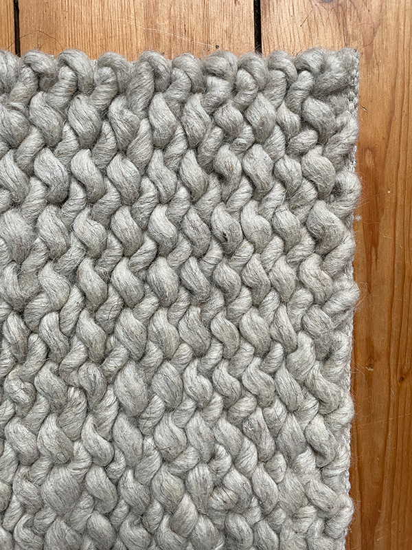 Close-up of a chunky knit light gray wool rug on a wooden floor.