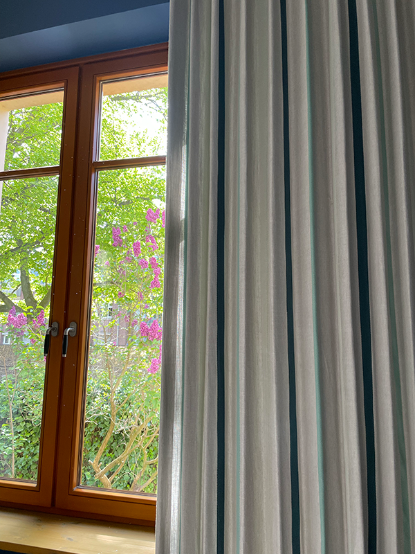 Wood-framed window partially covered by cream curtains with green vertical stripes, showing a garden with purple flowers and green foliage outside.