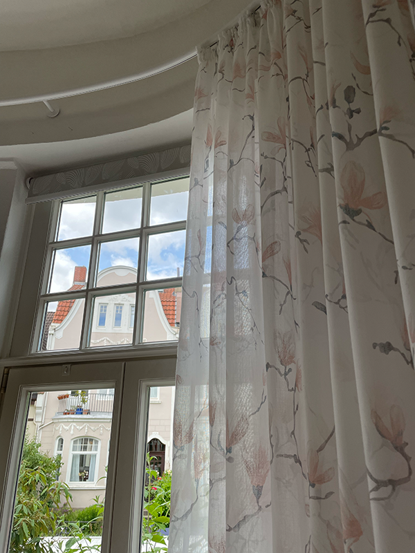 View through a window with sheer floral curtains partially drawn, showing a building with red tile roof and white facade outside.