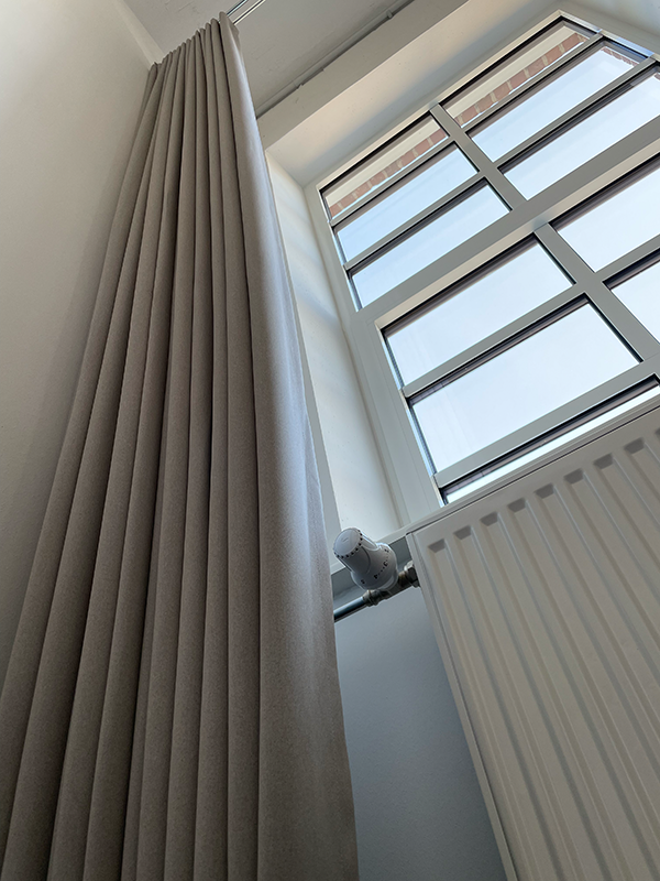 Tall beige curtain hanging next to a white radiator and a large window with grid-style panes.