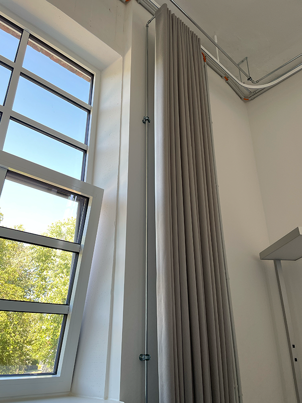 Tall window with multiple panes partially open, next to a floor-to-ceiling beige curtain and visible industrial ceiling pipes.