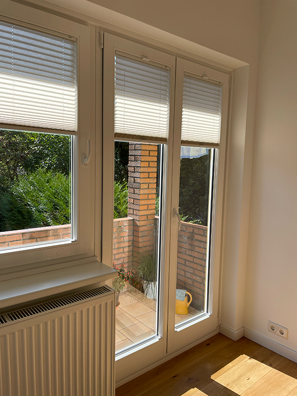 Bright room with white framed glass door and window partially covered by white blinds, a yellow watering can and plants visible outside on a tiled balcony.