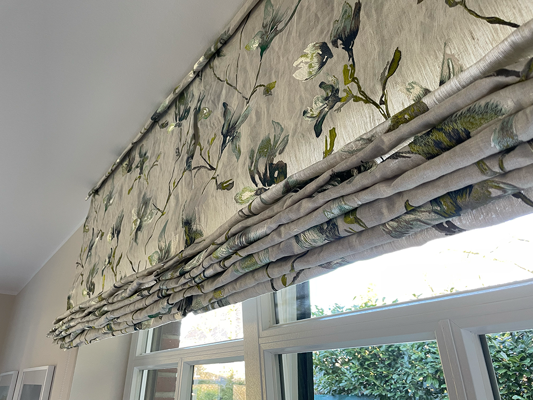 Close-up of a floral patterned Roman shade partially covering a window with outdoor greenery visible.