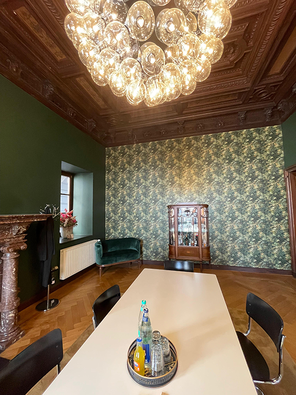 Elegant meeting room with a large white table, black chairs, decorative green wallpaper, a wooden cabinet, velvet green sofa, ornate wooden ceiling, and a modern chandelier.