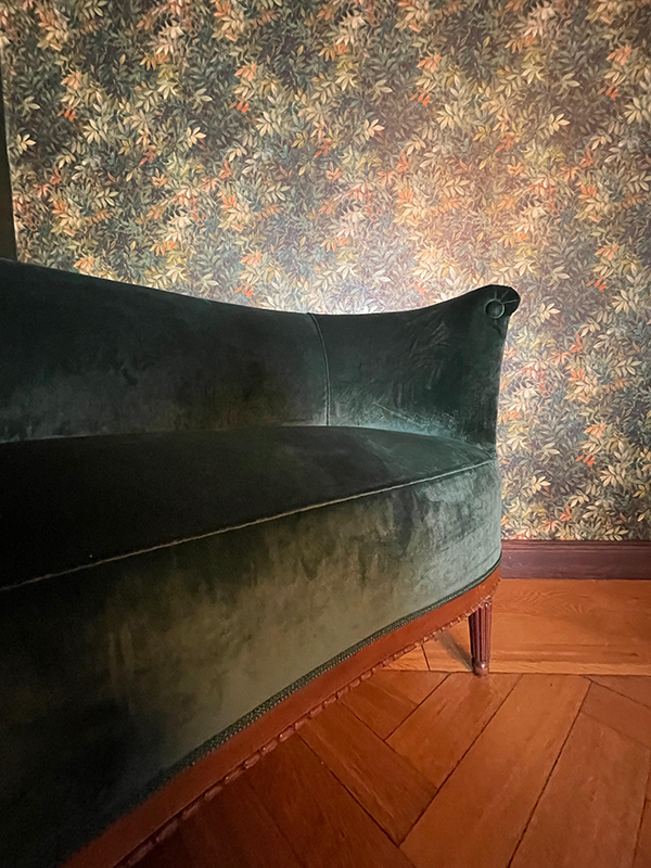 Curved dark green velvet sofa with wooden legs on a herringbone wood floor against floral patterned wallpaper.