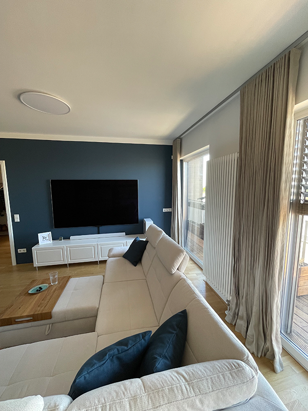 Modern living room with a large beige sectional sofa featuring navy blue cushions, a wooden coffee table, a flat-screen TV mounted on a dark blue accent wall, and large windows with beige curtains and white radiator.