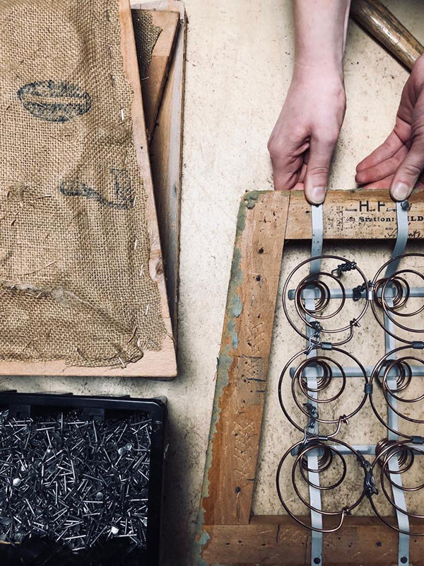 Hands working on an antique wooden frame with exposed coil springs and burlap fabric nearby, with a container of upholstery nails.